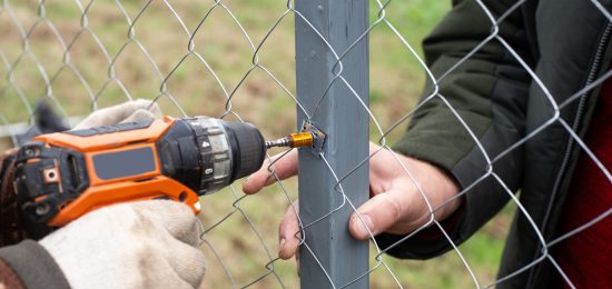 A man is building a fence at his dacha. Using an electric drill, he screws a metal mesh chain-link to the metal profile.