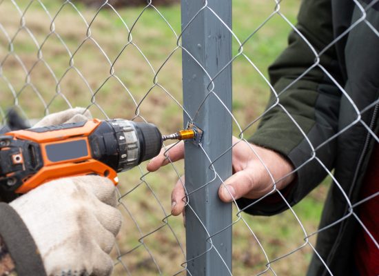 A man is building a fence at his dacha. Using an electric drill, he screws a metal mesh chain-link to the metal profile.