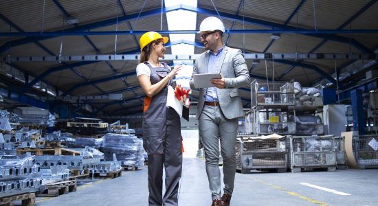 Manager supervisor and industrial worker in uniform walking in large metal factory hall and talking about increasing production.