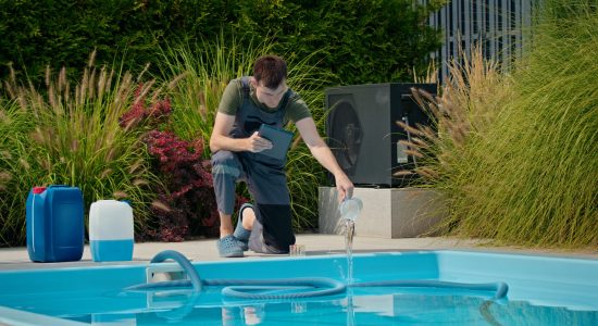 Pool technician kneeling by the swimming pool, pouring liquid while checking data on a tablet. Surrounded by greenery and chemical containers, emphasizing professional pool maintenance in an outdoor setting.