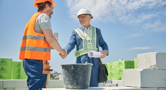 Smiling senior male contractor in eyeglasses and a bricklayer in a helmet greeting each other
