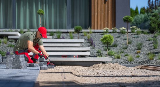 A worker is busy laying stone pavers in a beautifully designed outdoor space, surrounded by greenery on a bright afternoon.
