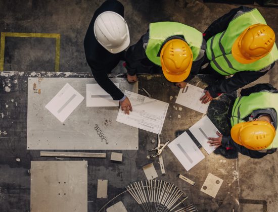 Workers and manager in safety helmets working with documents at factory